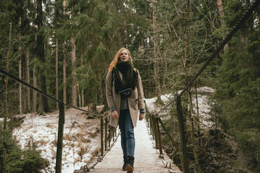 Woman in wool coat and scarf walking on footbridge in snowy woods - FSIF05079