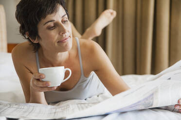 Frau entspannt sich auf dem Bett mit Kaffee und liest Zeitung - FSIF05131