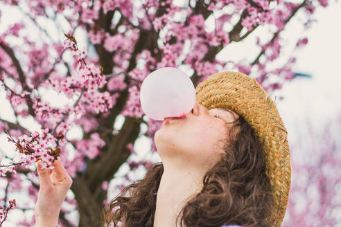 Junge romantische Teenager-Frau in Kleid und Hut macht Kaugummiblase gegen rosa Baum - ADSF05855