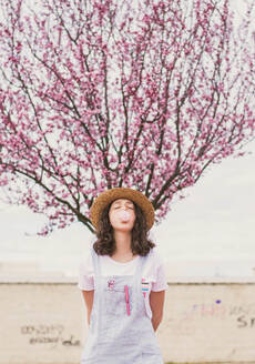 Junge romantische Teenager-Frau in Kleid und Hut macht Kaugummiblase gegen rosa Baum - ADSF05856