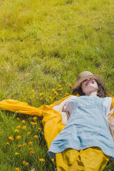 Junge stilvolle Frau in Regenmantel und Hut liegt auf einer grünen Wiese mit Blumen und träumt im Sonnenlicht - ADSF05861