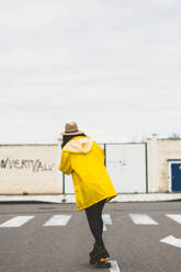 Junge schlanke Frau in gelbem Regenmantel fährt auf der Straße Skateboard - ADSF05867