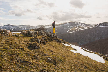 Seitenansicht eines Touristen, der auf einem Felsen an einem grünen Hang mit leichter Schneedecke vor malerischen Bergen in Wolken steht - ADSF05883