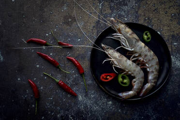 Small hot peppers lying near plate with delectable boiled shrimps on dark tabletop. - ADSF05911