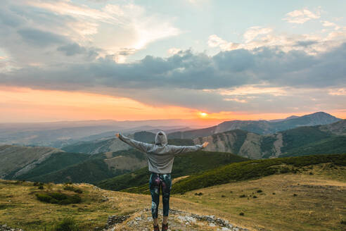 Rückenansicht einer sportlichen Frau im Kapuzenpulli mit erhobenen Armen vor dem Hintergrund einer majestätischen Berglandschaft im Sonnenuntergang - ADSF05924