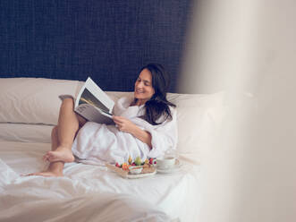 Smiling pretty woman lying on bed with served breakfast and reading magazine. - ADSF05926