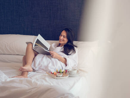 Lächelnde hübsche Frau, die auf dem Bett liegt, Frühstück serviert und eine Zeitschrift liest. - ADSF05926
