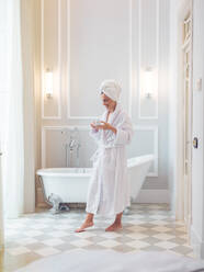 Cheerful pretty woman in bathrobe standing with plastic flask in bathroom. - ADSF05927