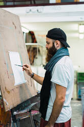 Side view of artist in cap and scarf standing at easel and starting to paint. - ADSF06023