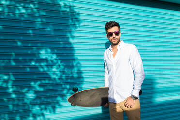 Young handsome brunet in casual and sunglasses posing with skateboard against turquois panel wall. - ADSF06039