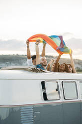 Gruppe von fröhlichen, unterschiedlichen Menschen, die in einem Retro-Van mit geöffnetem Dach stehen und eine LGBT-Flagge über den Köpfen halten, während sie gemeinsam in der Natur unterwegs sind - ADSF06055