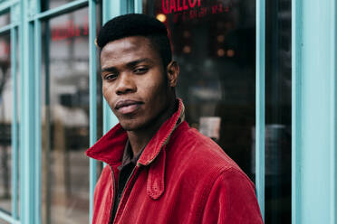 Stilvoller afrikanischer Mann, der seine Hände in die Taschen seines roten Pullovers steckt, steht am Eingang eines Cafés - ADSF06076