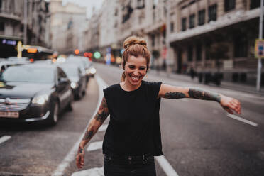 Porträt eines fröhlichen Mädchens mit tätowierten Armen, das mitten auf der Straße tanzt, während die Autos wegfahren.Bokeh - ADSF06102