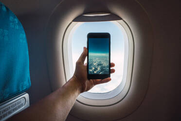 Crop Hand hält Smartphone und ein Bild von unendlichen weißen Wolken hinter dem Fenster des Flugzeugs - ADSF06107