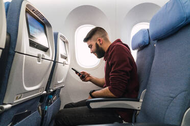 Seitenansicht eines gutaussehenden bärtigen Mannes, der auf einem bequemen Sitz in einem modernen Flugzeug sitzt und auf seinem Smartphone surft - ADSF06113