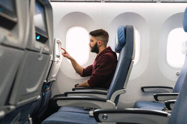 Side view of bearded guy browsing aircraft gadget while sitting on comfortable seat inside modern plane - ADSF06114