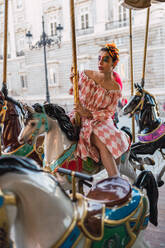 Attractive young female in theatrical costume looking away while sitting on horse of carousel on blurred background of amusement park - ADSF06135