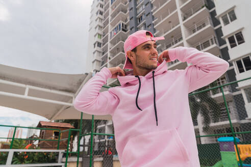 Gutaussehender junger Mann, der lächelt und wegschaut, während er auf dem Sportplatz steht und die Kapuze seines schicken rosa Hoodies zurechtrückt - ADSF06170
