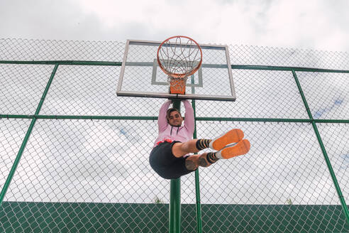 Von unten Schuss von fröhlichen jungen Mann Blick auf die Kamera, während hängen auf Basketball-Ring, während die Zeit auf Sportplatz - ADSF06171