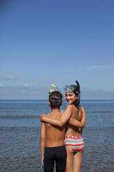 Bruder und Schwester tragen Schnorchel und umarmen sich am Strand - CAIF28389