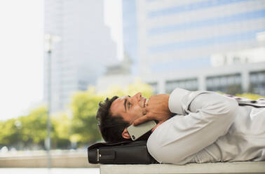 Businessman laying on briefcase and talking on cell phone in urban park - CAIF28434