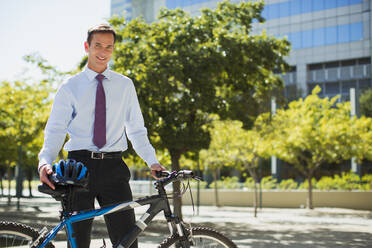 Smiling businessman with bicycle and helmet in urban park - CAIF28464