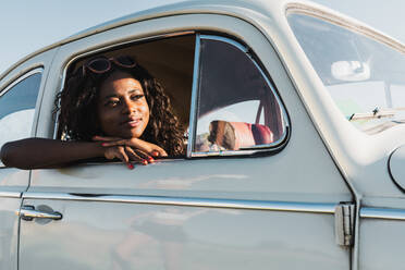 Happy young black woman in stylish sunglasses looking out of car window enjoying summer sunlight - ADSF06207