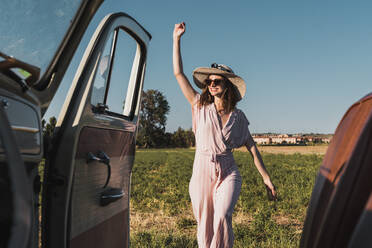 Blick aus dem Inneren eines Retro-Autos auf eine fröhliche, trendige Frau mit Hut und Sonnenbrille, die aufgeregt in der Natur steht und lacht - ADSF06212