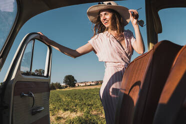 Blick aus dem Inneren eines Retro-Autos auf eine fröhliche, trendige Frau mit Hut und Sonnenbrille, die aufgeregt in der Natur steht und lacht - ADSF06213