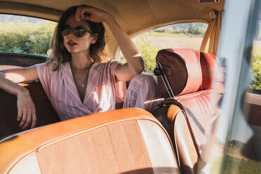 Beautiful confident woman in pink and sunglasses sitting on back seat in retro car looking away in sunlight - ADSF06215