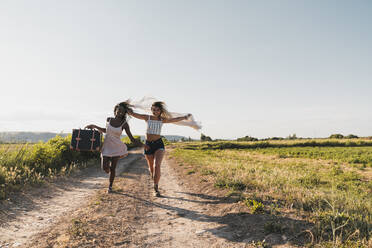 Fröhliche stilvolle multiethnische Frauen mit Koffer und Schal laufen aufgeregt auf der Straße im Sommer grüne Landschaft - ADSF06216
