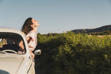 Glückliche junge Frau mit modischer Sonnenbrille, die aus dem Autofenster schaut und die Sommersonne vor grünen Bäumen und blauem Himmel genießt - ADSF06219