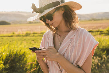 Brunette in pink clothes and elegant hat surfing smartphone on background of green summer countryside in?sunlight - ADSF06220