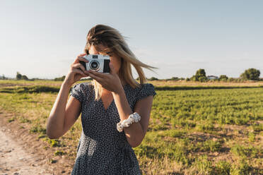 Frau im Kleid, die ein Foto mit einer Retro-Kamera auf dem Hintergrund einer grünen Sommerlandschaft im Sonnenlicht macht - ADSF06222