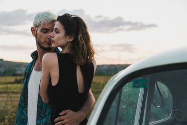 Embracing man and woman in elegant dress standing near car on background of landscape - ADSF06226