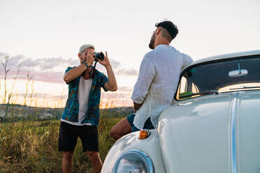 Lässiger Mann im Hemd, der eine Fotokamera benutzt und einen Mann mit Auto in einer sommerlichen Landschaft fotografiert - ADSF06231