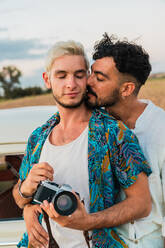 Küssende verliebte Männer, die sich zärtlich umarmen, während sie mit einer Fotokamera in der Natur stehen und das Reisen genießen - ADSF06234