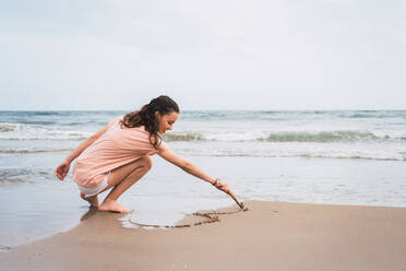 Junges hübsches Mädchen hockt und malt mit einem Stock auf Sand am Meeresufer - ADSF06238