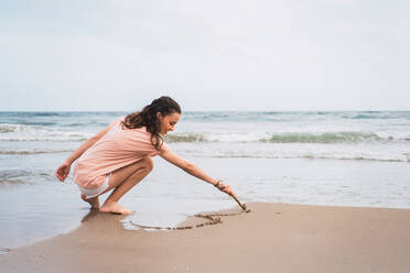 Young pretty girl squatting and painting with stick on sand on seashore - ADSF06238