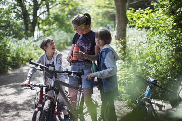 Mutter und Söhne trinken Wasser bei einer Fahrradtour im sonnigen Park - CAIF28574