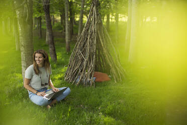 Smiling woman using laptop outside branch teepee in woods - CAIF28594