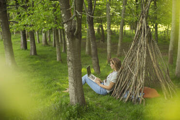 Woman with laptop relaxing at branch teepee in woods - CAIF28599