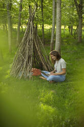 Frau mit Laptop entspannt sich am Zweig-Tipi im Wald - CAIF28611