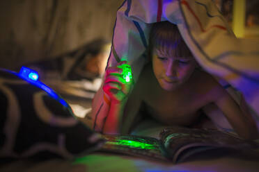 Junge mit grüner Taschenlampe, der unter einer Decke eine Zeitschrift liest - CAIF28775