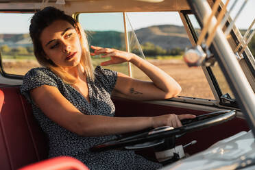 Junge Frau sitzt auf dem Fahrersitz und steuert einen Oldtimer-Van, während sie an einem sonnigen Tag auf dem Land unterwegs ist - ADSF06289