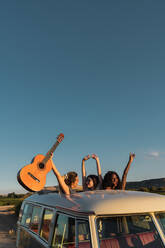 Three cheerful young ladies holding guitar and having fun while standing in hatch in roof of retro van in nature - ADSF06291