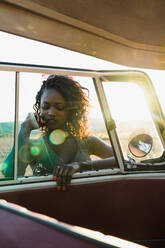 Schöne afroamerikanische Frau lächelt und schaut durch das Fenster eines Oldtimers in die Kamera, während sie an einem sonnigen Tag Zeit in der Natur verbringt - ADSF06292