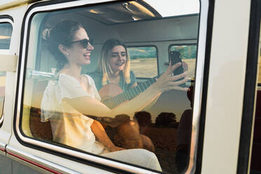 Women playing acoustic guitar and laughing while sitting together inside retro van during trip - ADSF06293