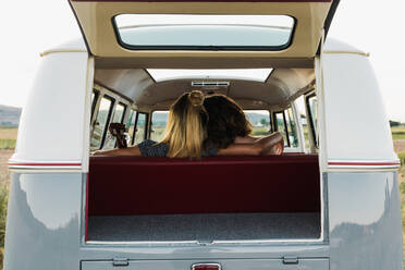 Back view of two women embracing each other while sitting on back seat of retro van in nature - ADSF06294