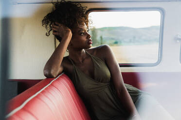 Attractive African-American female sitting on comfortable back seat of vintage van and looking away while traveling in countryside - ADSF06295